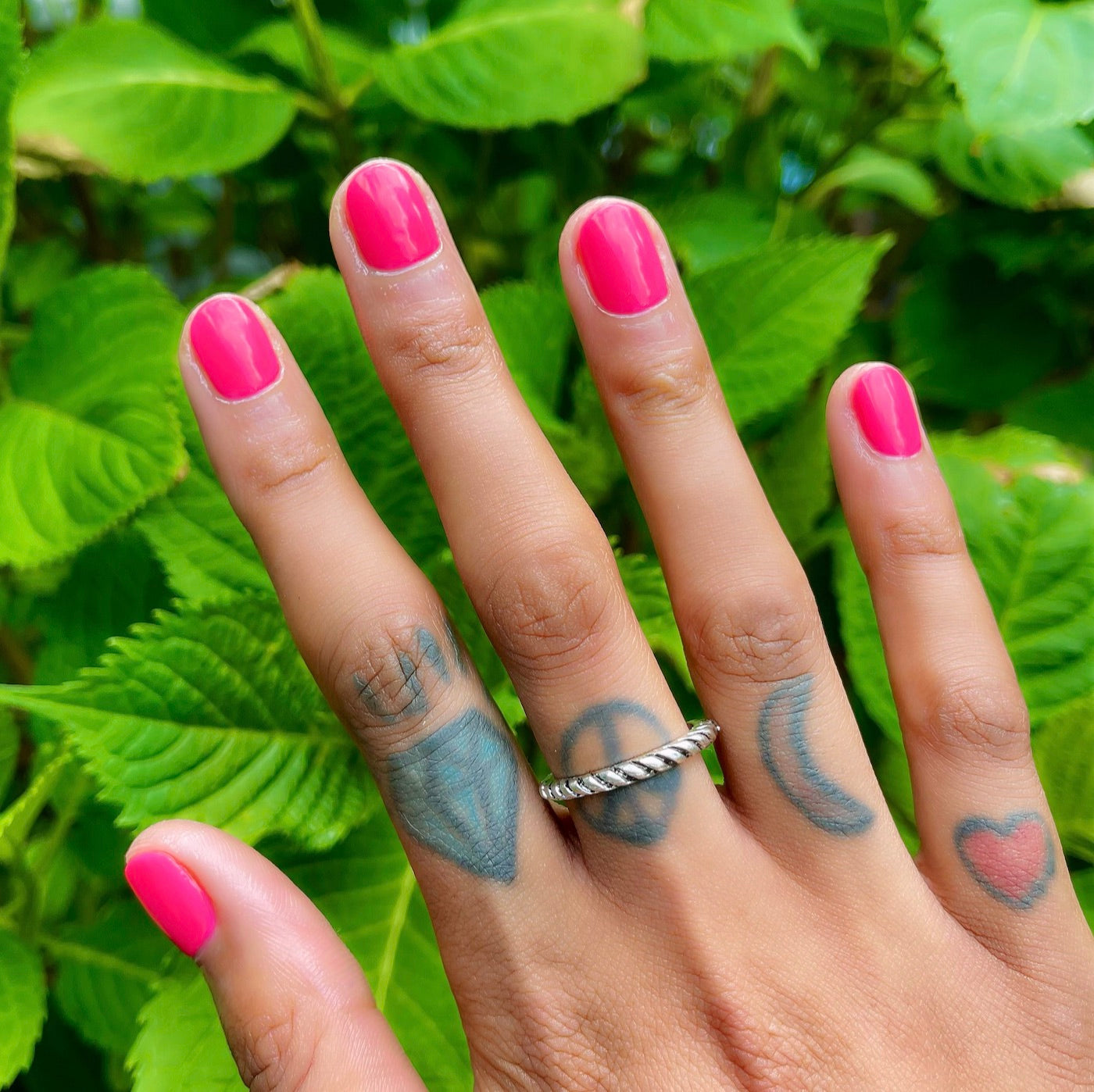 Hand with vibrant Hot Strawberry polish in bright strawberry pink against green leafy background for at-home mani