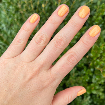 Hand with nails painted in bright cantaloupe orange Counselor In Training gel polish against natural green background