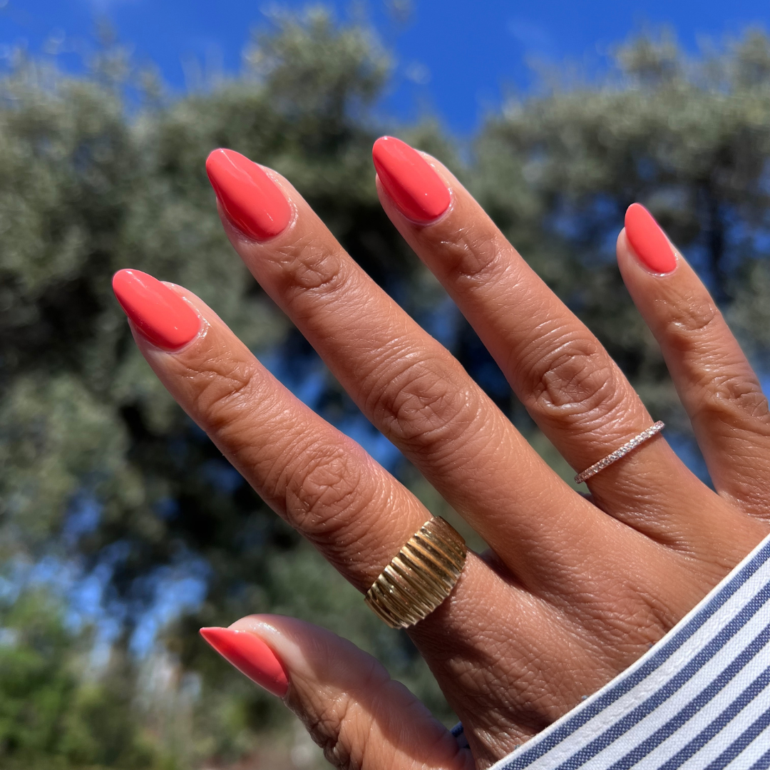 Hand with almond-shaped nails painted in Cape showing a vibrant pinky-red manicure with outdoor trees and blue sky background