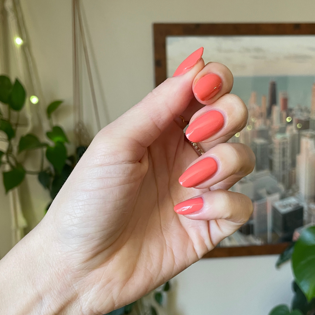 Hand with oval nails painted in Cape showing a glossy preppy pinky-red manicure with a city skyline blurred background