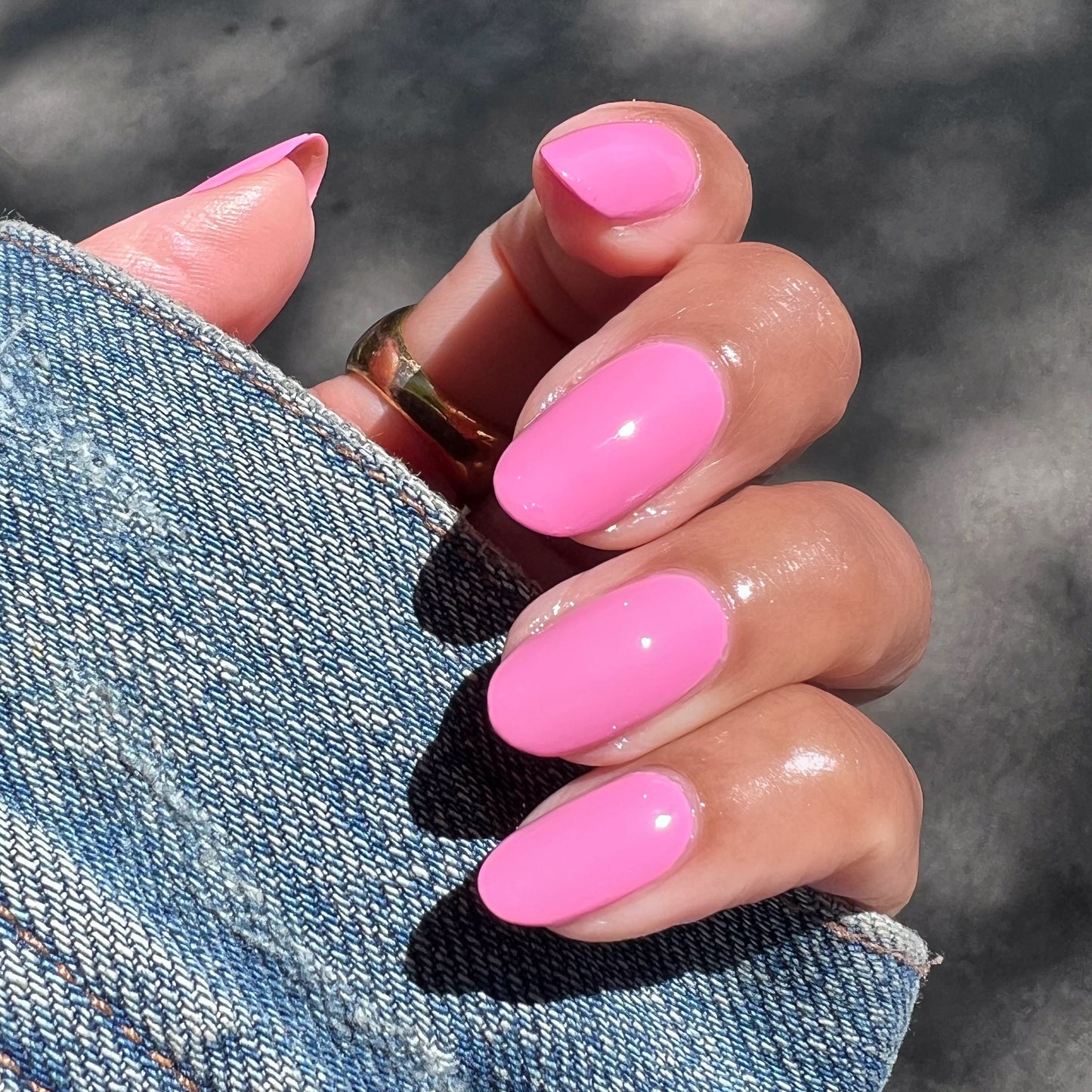 Hand with glossy bright pink Taffy gel polish on nails holding denim jacket sleeve in natural sunlight manicure view