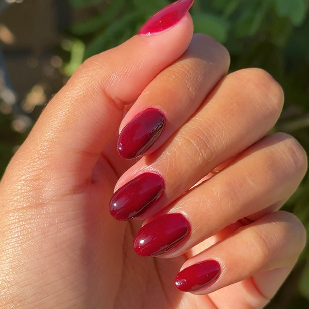 Hand with glossy deep red gel polish nails held against a blurred green outdoor background
