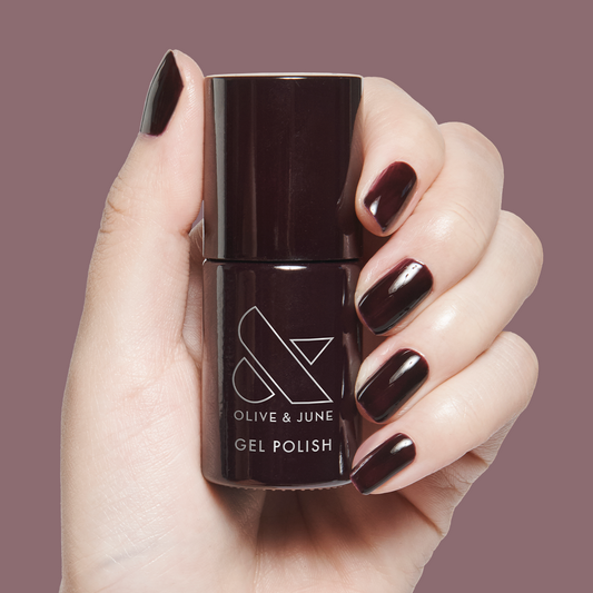 Hand holding bottle of Covet - Gel gel polish with nails painted in a deep dark plum shade on muted mauve background
