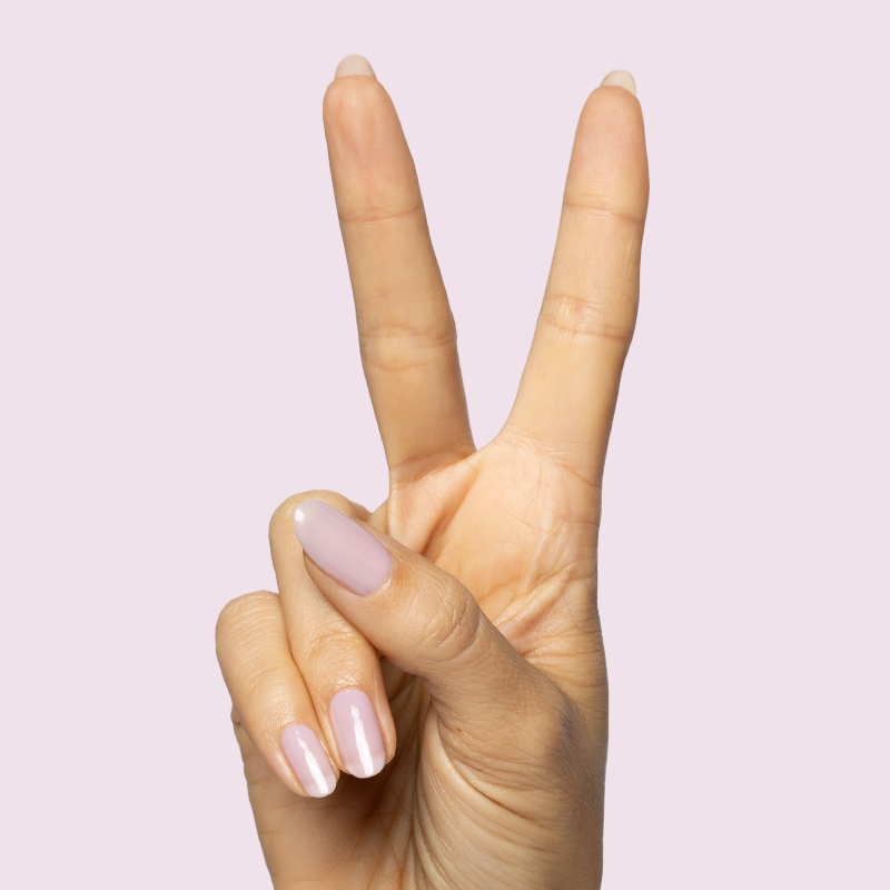 Hand with natural nails painted in sheer milky pastel lavender Dreamy gel polish on soft pink background showing peace sign