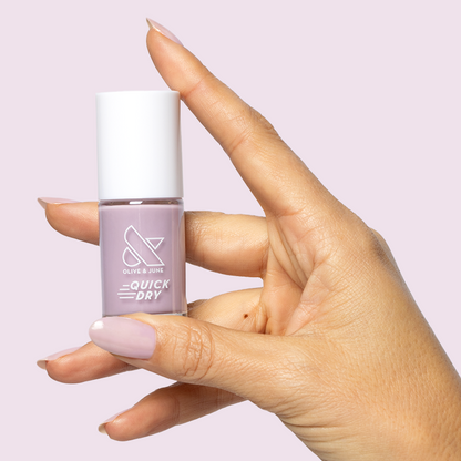 Hand holding a bottle of Dreamy sheer milky pastel lavender quick dry gel polish against a soft lavender background