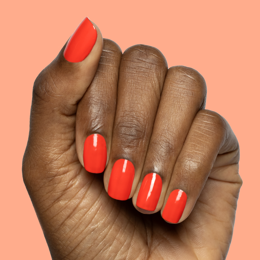 Hand with short nails painted in bright orange-red Snapdragon quick dry polish on soft coral background