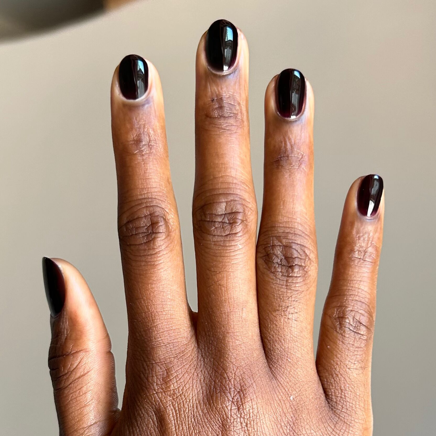 Hand with dark skin tone showing glossy nails painted in Covet - Gel, a deep cool plum shade, on a light background