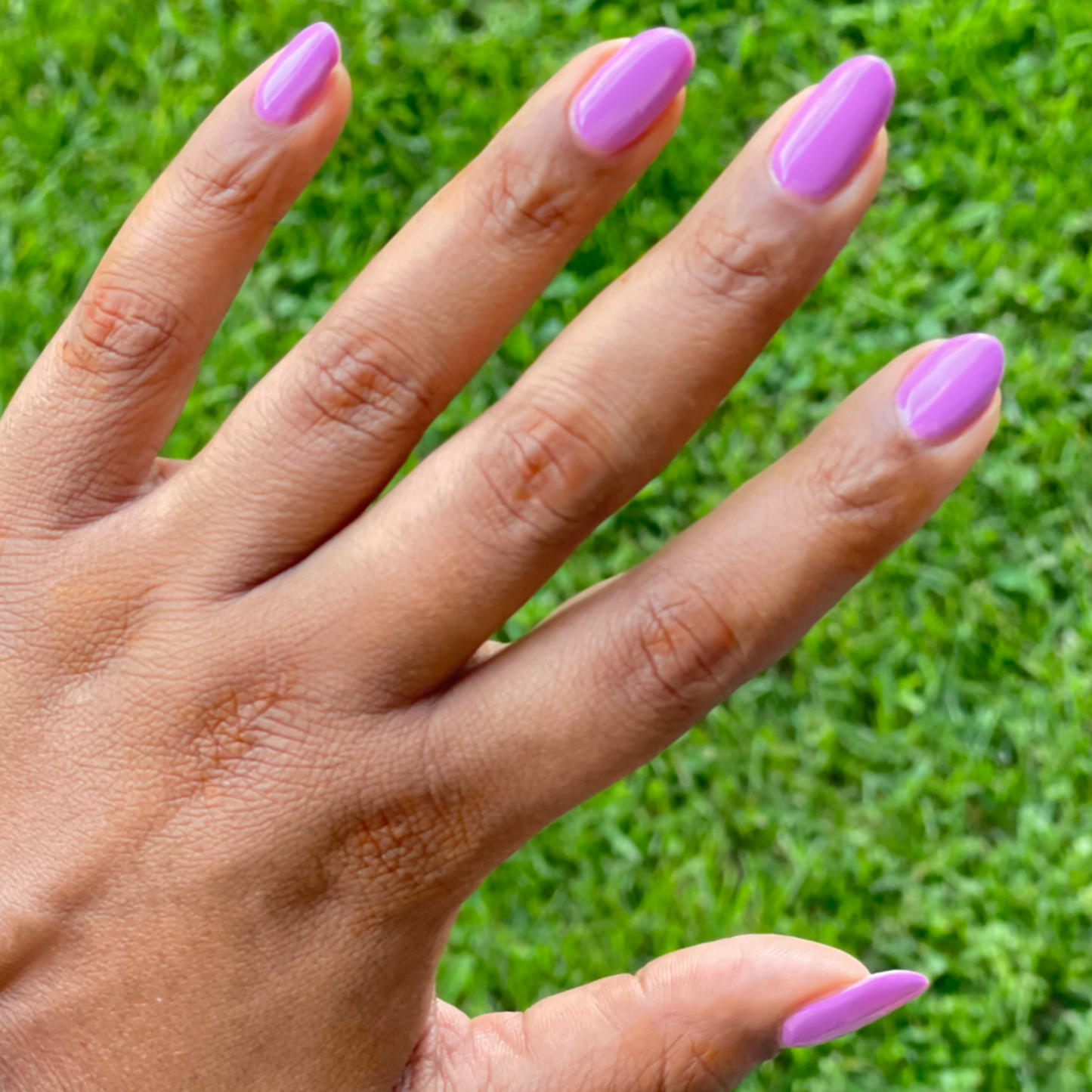 Hand with nails painted in bright floral purple pink shade using Petal - Gel against green grass background