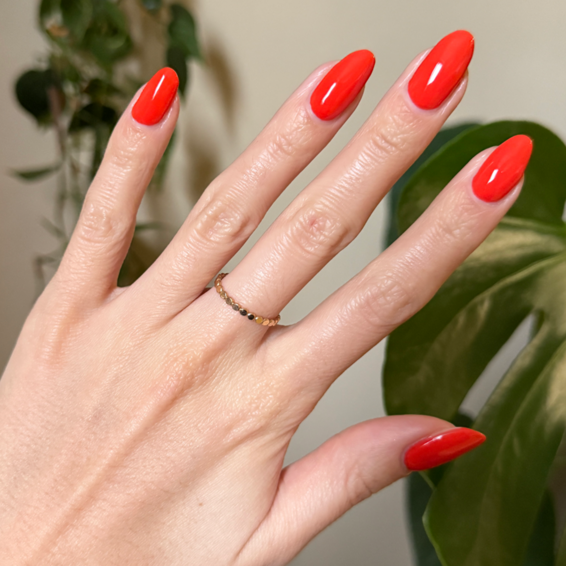 Hand with almond-shaped nails painted in bright Snapdragon orange-red gel polish against green plant background
