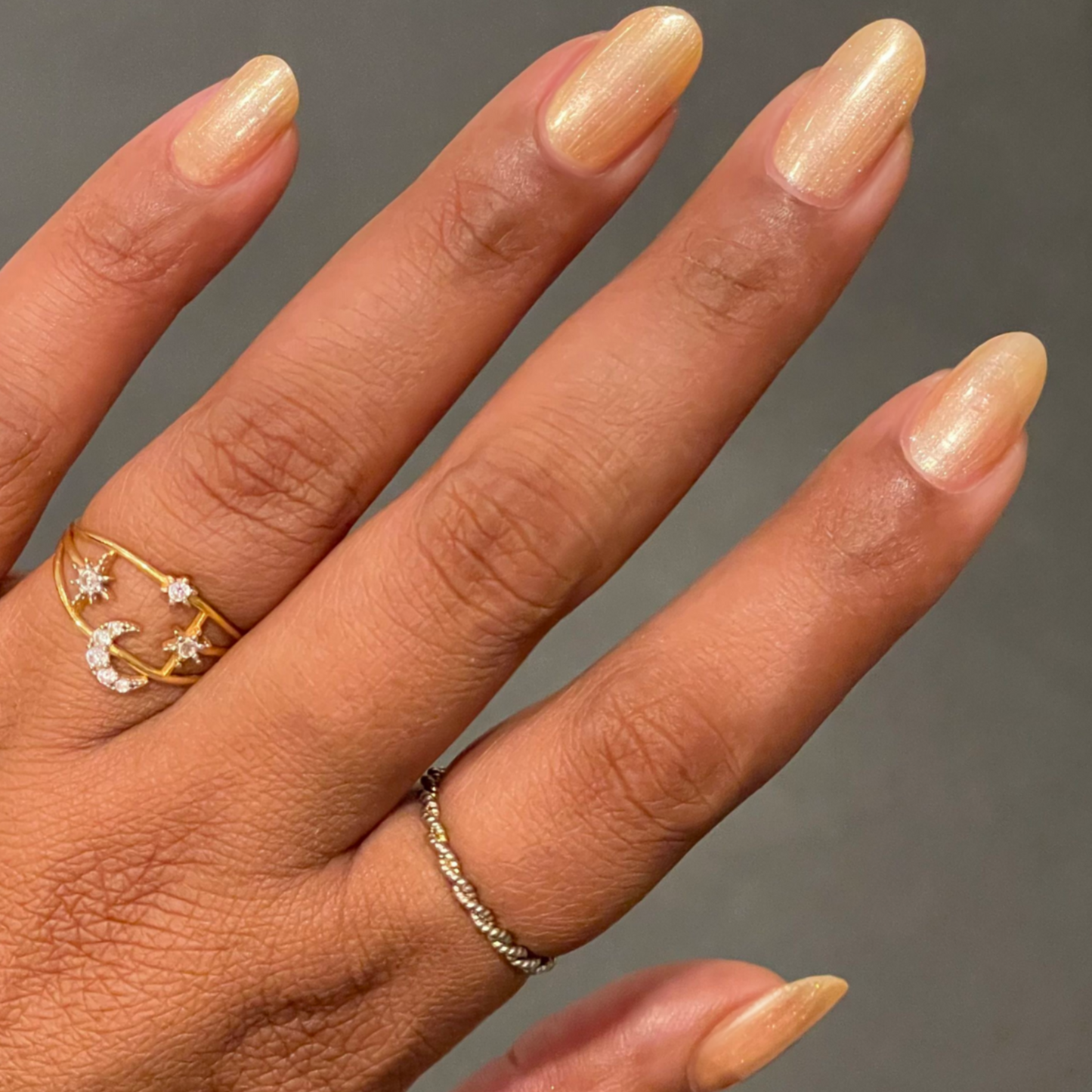 Hand with medium-length natural nails polished in Sunlit warm golden peach shimmer gel polish with delicate rings